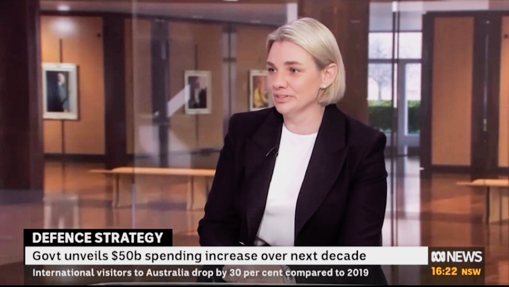 Professionally dressed woman talking at a news desk during a televised news program. 