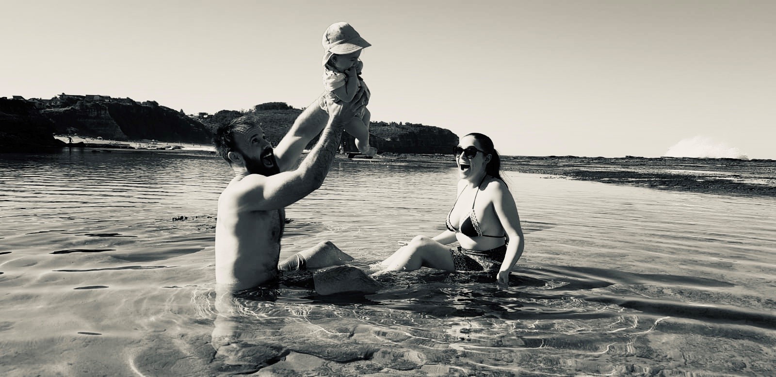 Young family consisting of a man and women in swimmers sitting in shallow water at the beach play with their toddler.