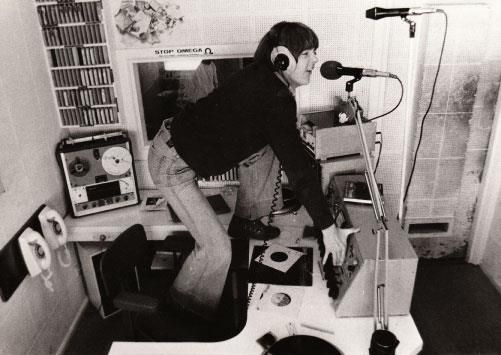 A black-and-white photo from the 1970s showing a radio booth in which an announcer is standing on their chair and speaking into the microphone.